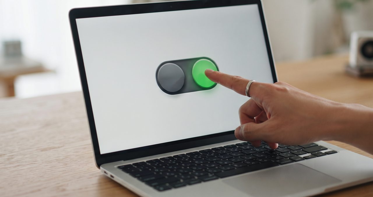 手元のクローズアップで、指がノートパソコンの画面いっぱいに大きく表示されたトグルスイッチをグレーから緑色へ切り替えている瞬間