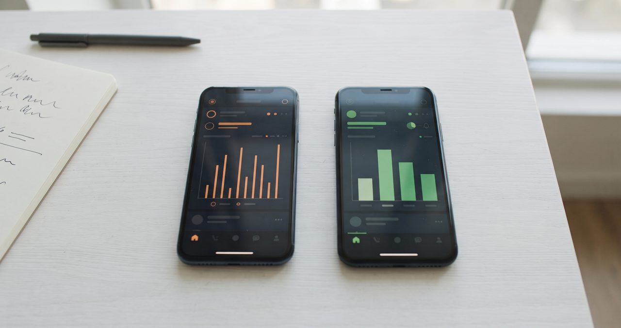 白い机の上に2台のスマートフォンが並べて置かれ、それぞれ異なる色の棒グラフ風の抽象的なグラフが画面に表示されている真上からのフラットレイ