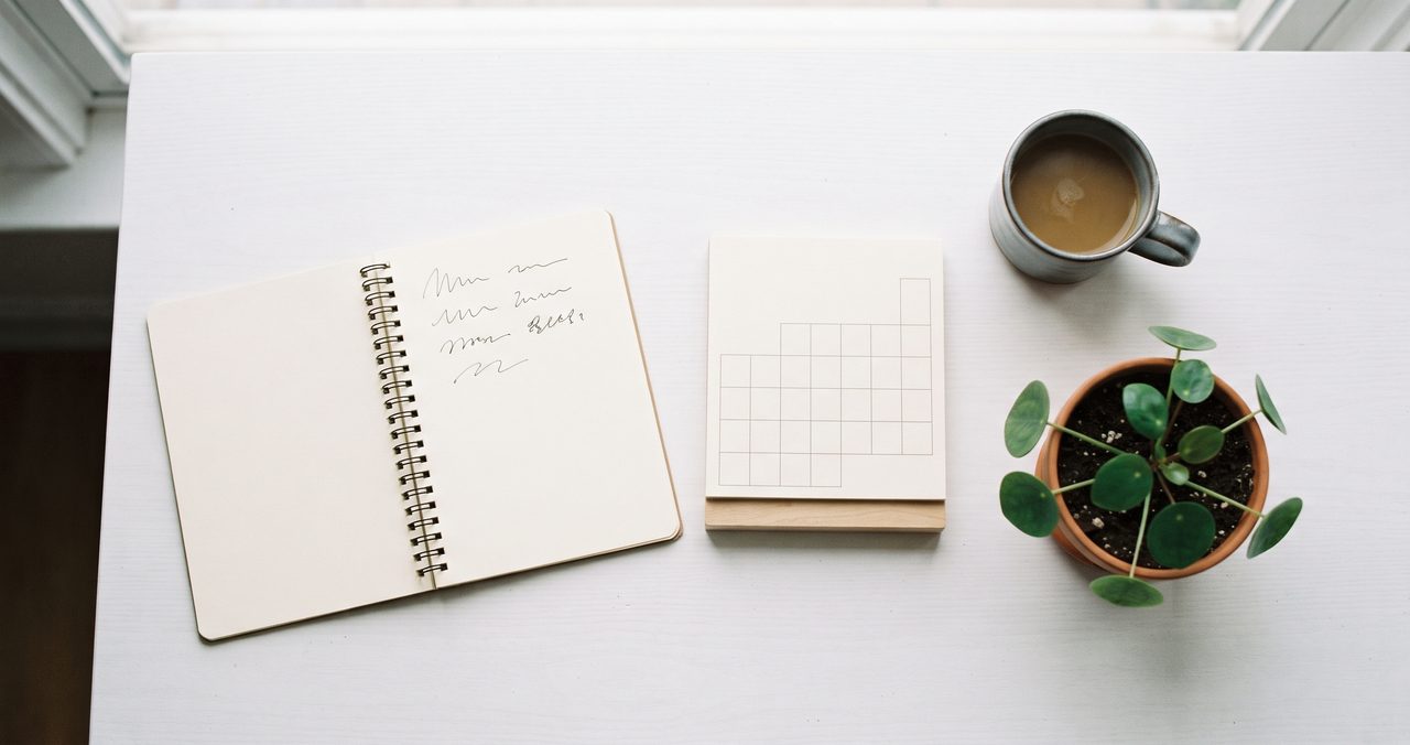 白い机を真上から見たフラットレイ、左に手書きの波線が書かれた開いたノート、中央に木製スタンドに立てられた方眼(マス目)のメモパッド、右にコーヒーの入ったマグカップと観葉植物の小さな鉢が並んでいる