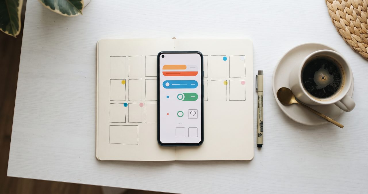 手描きの週間カレンダーのノートの上にスマートフォンが置かれ、画面に抽象的なトグルアイコンが表示されている真上からのフラットレイ、隣にコーヒーカップ